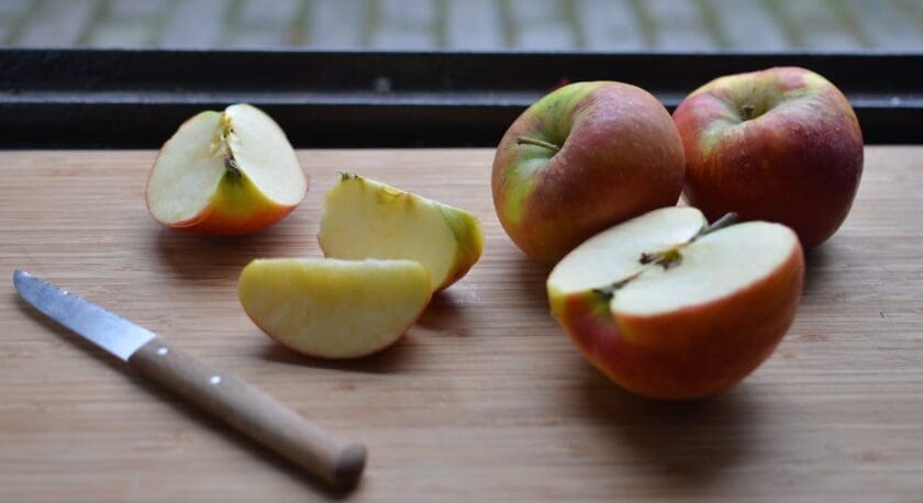 Two whole apples and several apple slices are arranged on a wooden cutting board, accompanied by a small knife with a wooden handle. The apples have a red and green skin, and the slices reveal the pale yellow flesh inside.