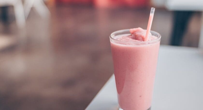 A tall glass filled with a pink smoothie, with a matching pink straw, placed on a white surface. The background is blurred, highlighting the smoothie.