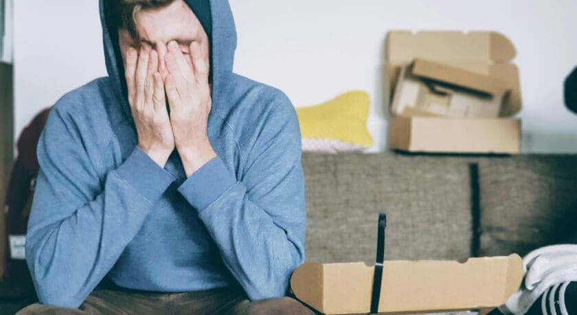 A person wearing a blue hoodie and a black beanie is sitting on a couch with their face buried in their hands, appearing distressed. In front of them are several small boxes, some open, and a glass cup. The background shows a gray couch with a yellow pillow and some cardboard boxes.