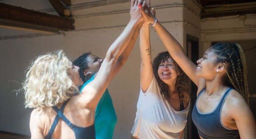 Four women in athletic wear are standing in a circle indoors, smiling and giving each other a high-five with their arms raised. The setting appears to be a gym or studio with wooden beams and a neutral-colored wall in the background.