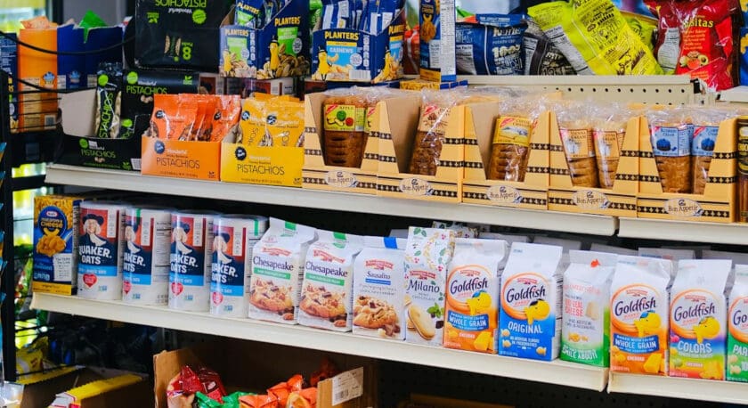 Shelves stocked with various packaged snacks including Wonderful pistachios, Planters nuts, Bon Appetit pastries, Quaker Oats, Chesapeake cookies, Pepperidge Farm Sausalito and Milano cookies, Goldfish crackers in multiple varieties, Kraft macaroni and cheese, and assorted candy like Skittles and Cheetos in boxes below.