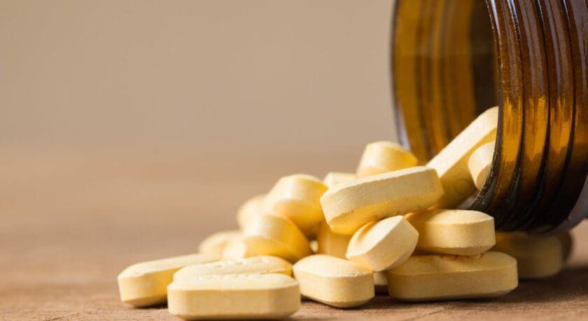 Yellow oval tablets spilling out of a brown glass bottle onto a wooden surface.