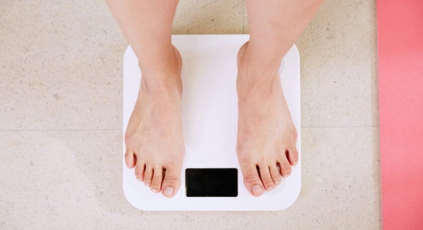 Bare feet standing on a white digital bathroom scale with a black display screen, placed on a light-colored tiled floor next to a pink mat.