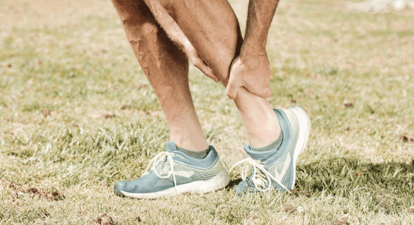 A person wearing blue and white running shoes with yellow laces is standing on grass, holding their right ankle with both hands, suggesting an ankle injury or pain.