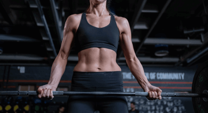 A muscular woman in a black sports bra and black leggings is gripping a barbell, preparing to lift it in a gym setting. Her toned arms and abs are visible, and she has red nail polish on her fingernails. The background shows gym equipment and a wall with partially visible text.