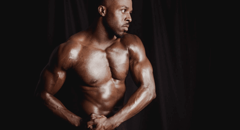 Muscular shirtless man with dark skin posing against a black background, showing defined chest, arm, and shoulder muscles. He is looking to the side with his hands clasped in front of him.
