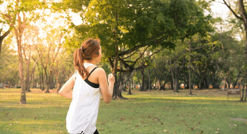 A woman jogging in a park during the late afternoon, wearing a white sleeveless top and black shorts, with her hair tied back in a ponytail. The park is filled with green grass and trees, with sunlight filtering through the foliage.