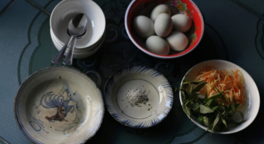 A collection of bowls and plates on a dark green table, including a red bowl with white eggs, a white bowl with shredded vegetables and green leaves, a small plate with salt and pepper, a decorative plate with a rooster design, and a stack of white bowls with two spoons on top.