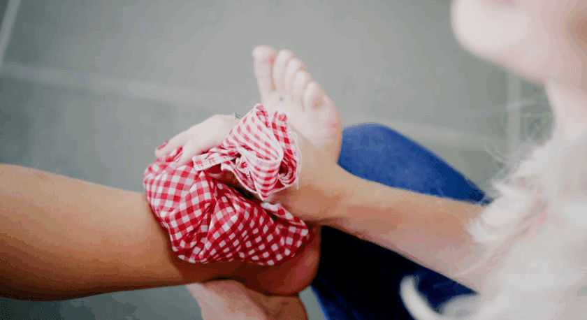 A person is holding another person's foot and applying a red and white checkered cloth, possibly an ice pack or compress, to the ankle area. The background is blurred, focusing on the foot and hands.