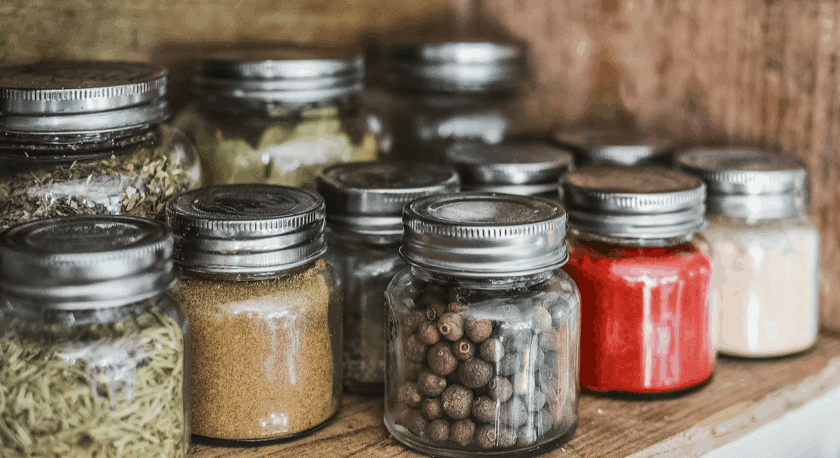 Small glass jars with metal lids filled with various spices and herbs, arranged on a wooden shelf. The spices include whole peppercorns, ground powders, dried herbs, and bright red and white powders.