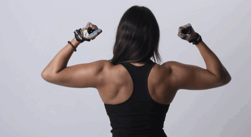 A person with long dark hair wearing a black sleeveless athletic top and colorful workout gloves is flexing their arm muscles, showing their back against a plain light gray background.