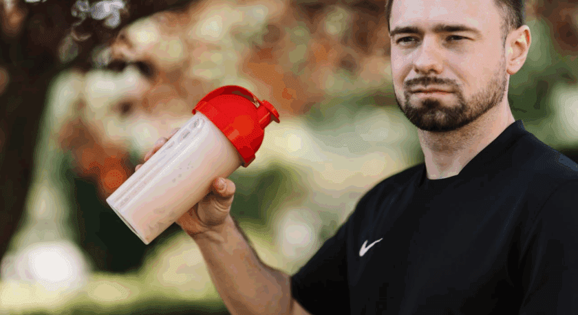 Man in a black Nike shirt holding a beige protein shaker bottle with a red lid outdoors.
