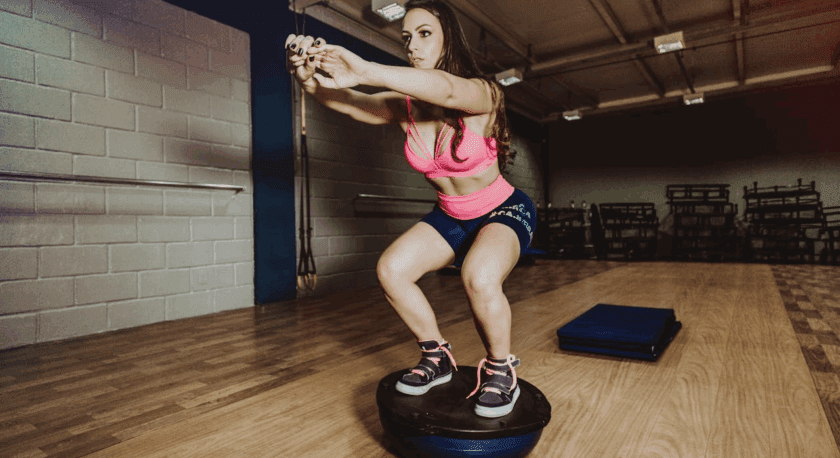 A woman in a pink sports bra and navy shorts is performing a squat exercise on a blue and black balance trainer in a gym with wooden flooring and gray brick walls.