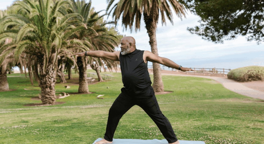 A man with a gray beard wearing a black sleeveless shirt and black pants is practicing yoga on a light blue mat outdoors. He is in a warrior pose with arms extended, standing barefoot on green grass with palm trees and a pathway in the background.
