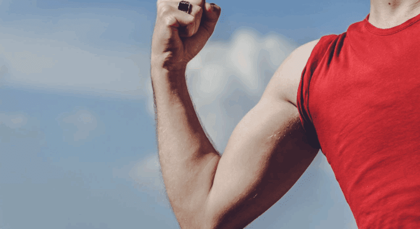 A person wearing a red sleeveless shirt flexing their right arm, showing a muscular bicep against a blue sky background. The person is also wearing a black ring on their right hand.