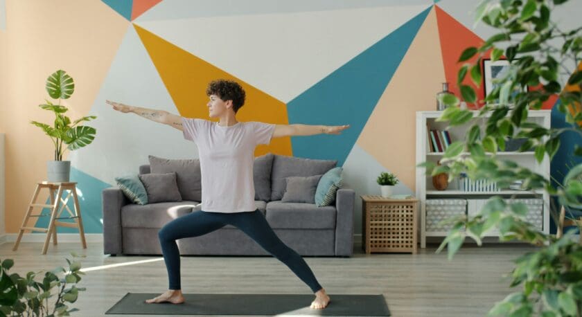 A person with short curly hair wearing a light pink t-shirt and dark blue leggings is practicing yoga in a brightly lit room. They are standing on a black yoga mat in a warrior pose with arms extended. The background features a gray sofa with cushions, a colorful geometric wall, a wooden stool with a potted plant, and a white shelving unit with books and decorative items. Green plants are visible in the foreground and background.